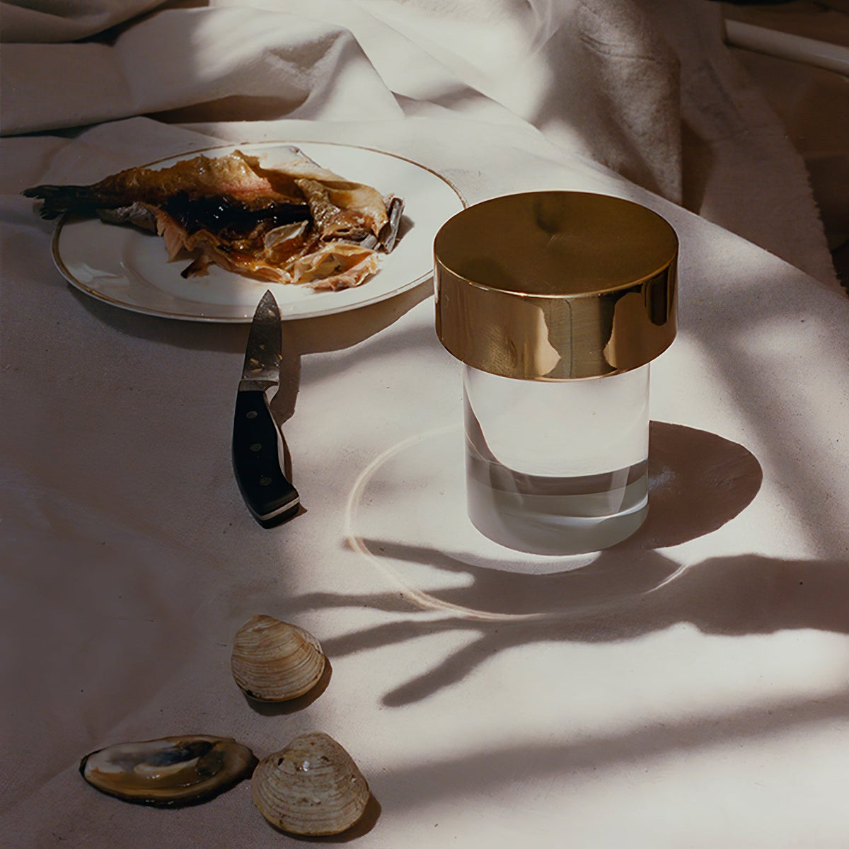 A large gold-capped Clérachic lamp on a white cloth beside a plate of fish, a knife and a few shells, with long shadows.