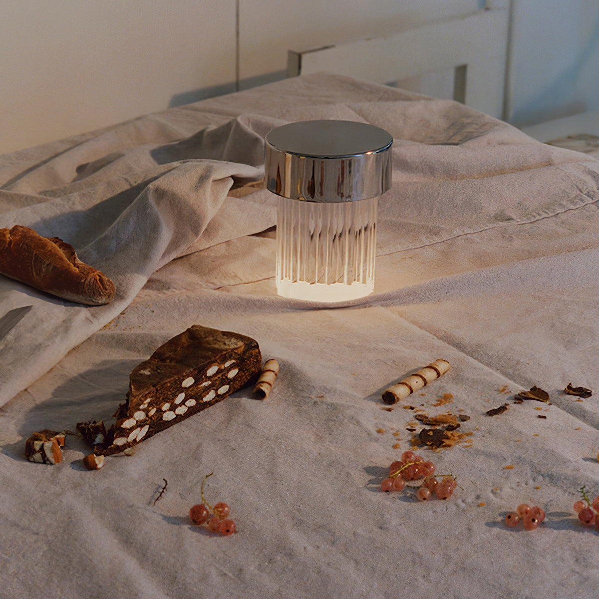 A fluted-glass Clérachic lamp with a chrome cap glowing on a rumpled bed, with bread and chocolate pieces scattered beside it.