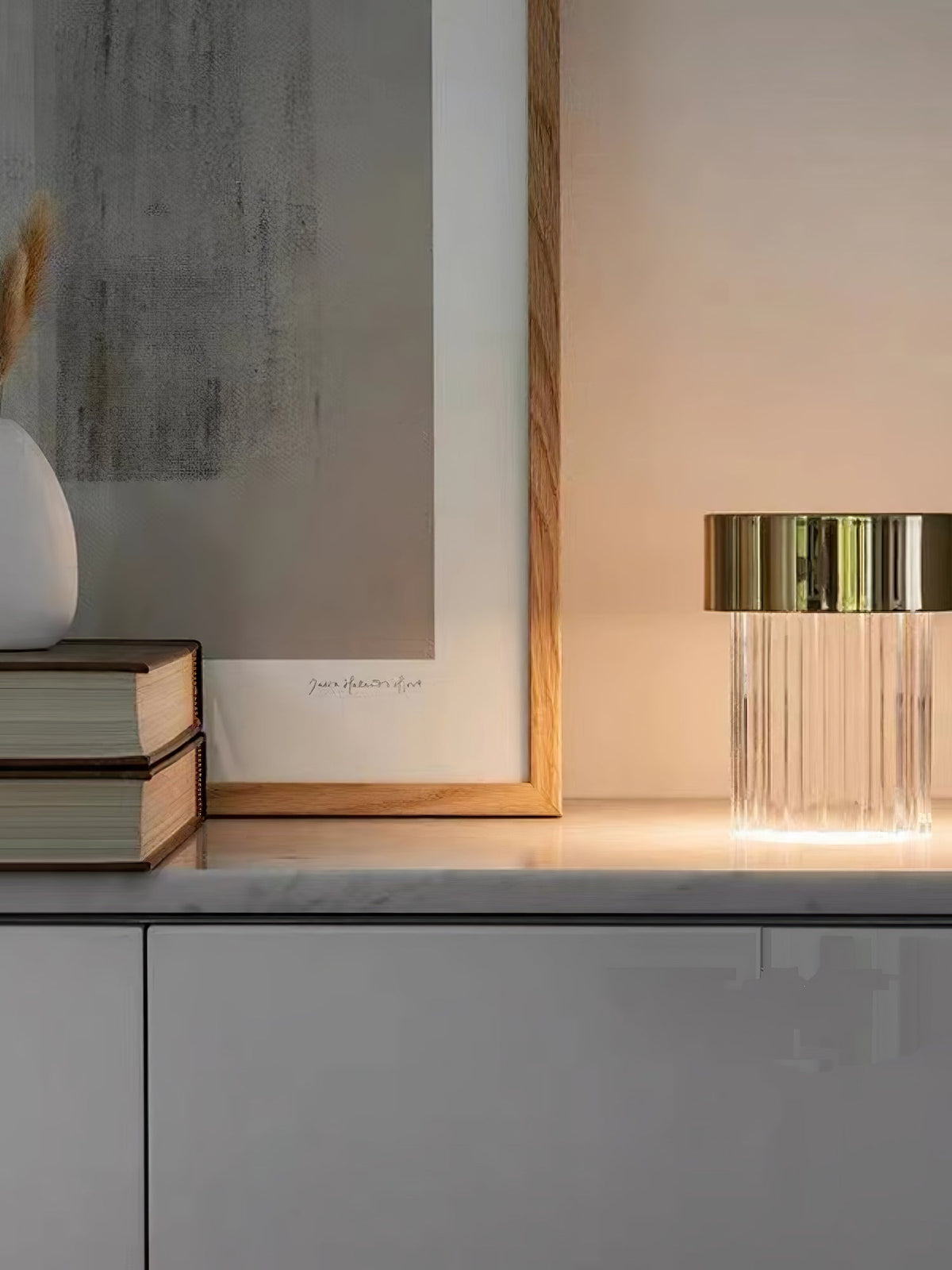 A fluted Clérachic lamp with a gold cap on a marble-topped sideboard beside stacked books and a pale framed print.