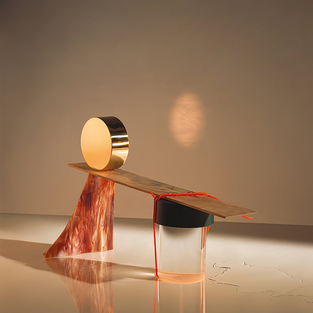 A Clérachic lamp balanced under a wooden plank carrying a gold disc, set up as a sculptural still life with an orange cord.