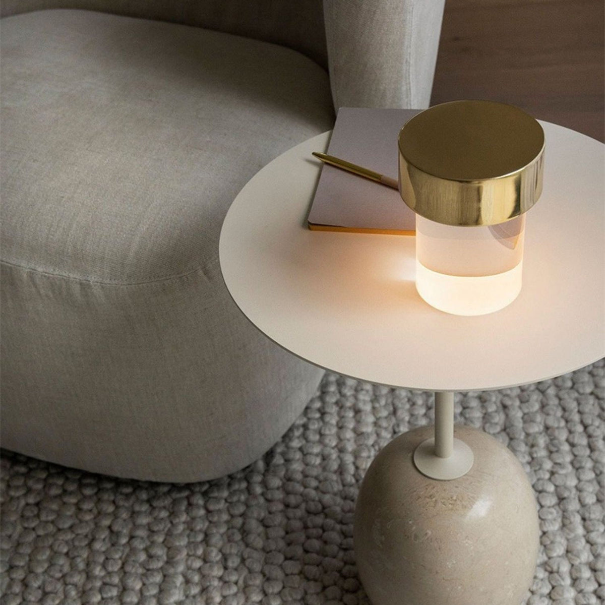 Looking down on a Clérachic lamp with a gold cap glowing on a small round side table by a cream sofa, with a notebook beside it.