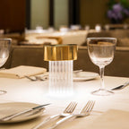 Close view of a fluted Clérachic lamp with a gold cap on a laid restaurant table, with glasses, plates and forks around it.