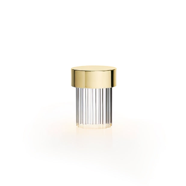 A Clérachic lamp with a gold cap and ribbed clear glass on white, with a soft warm glow beneath it.