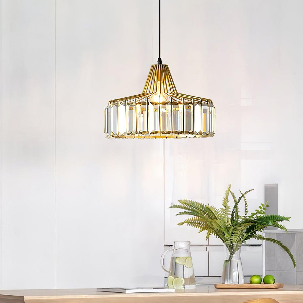 A gold-framed Clérachic crystal drum pendant hangs in a bright kitchen, with a fern and a jug of lemon water on the counter below.