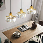 Three gold-framed crystal drum pendants from Clérachic at staggered heights over a wooden table with an open magazine and stacked plates.