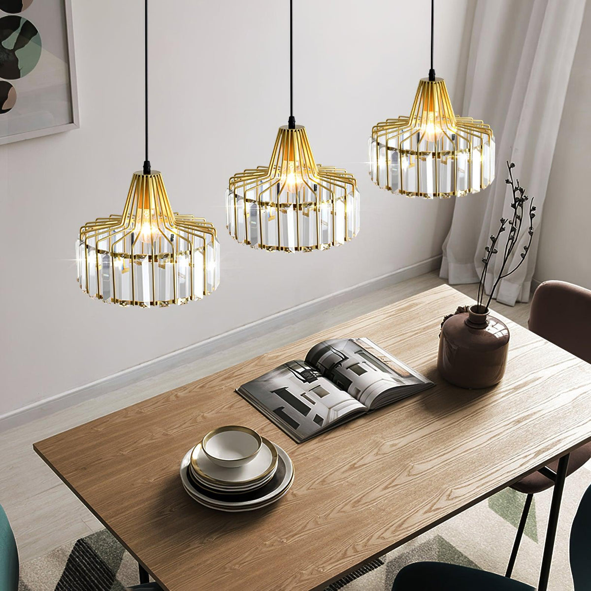 Three gold-framed crystal drum pendants from Clérachic at staggered heights over a wooden table with an open magazine and stacked plates.