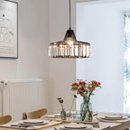 A black Clérachic crystal drum pendant, lit, hangs above a laid dining table with fresh flowers and a radiator in a bright room.