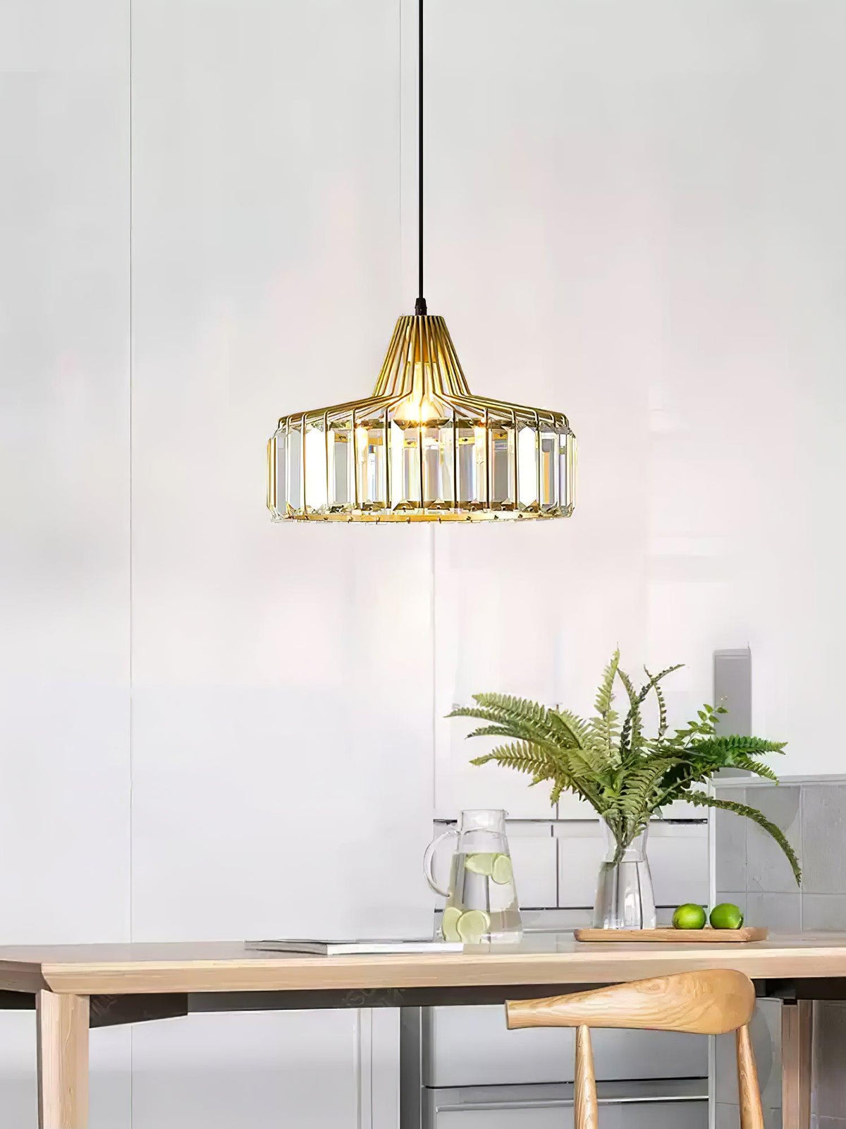 Gold crystal drum pendant from Clérachic hanging in a pale room above a wooden table, with a fern and a jug on the counter behind.