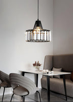 Small black-framed crystal drum pendant from Clérachic above a white table, with a grey sofa and moulded chairs in a neutral room.