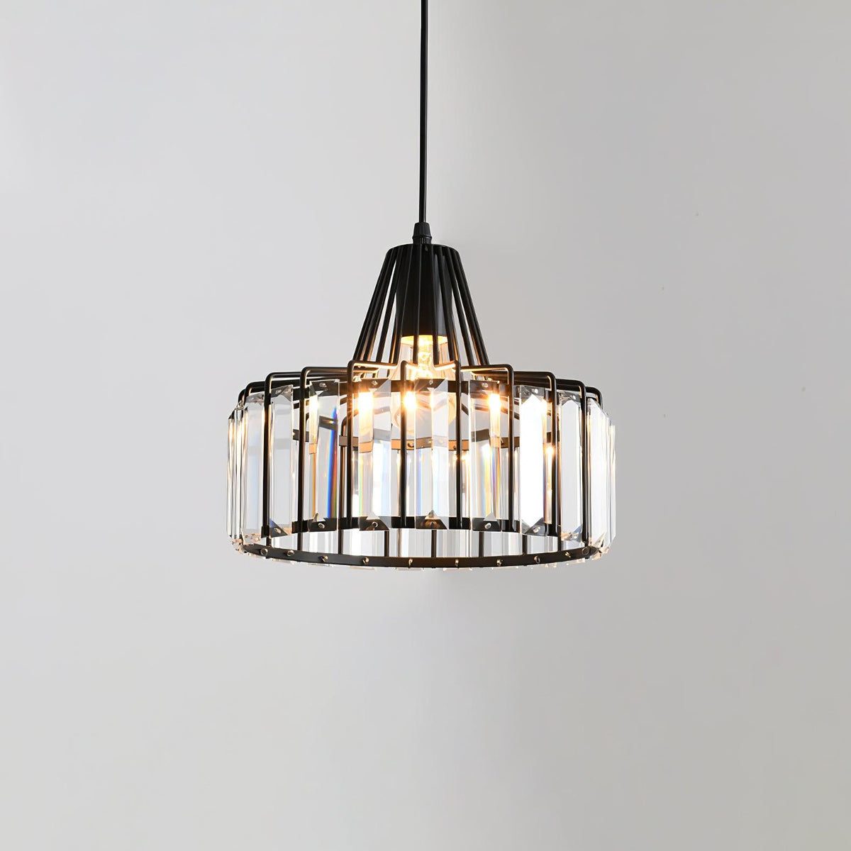 Black-framed Clérachic pendant with tall clear crystal prisms around the drum, lit from within, against a pale grey wall.
