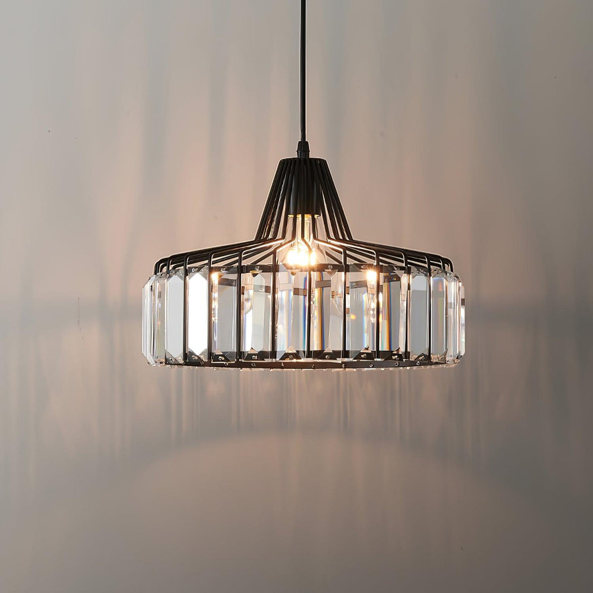 Lit black wire-framed crystal drum pendant from Clérachic, casting soft reflections on a warm grey wall behind it.