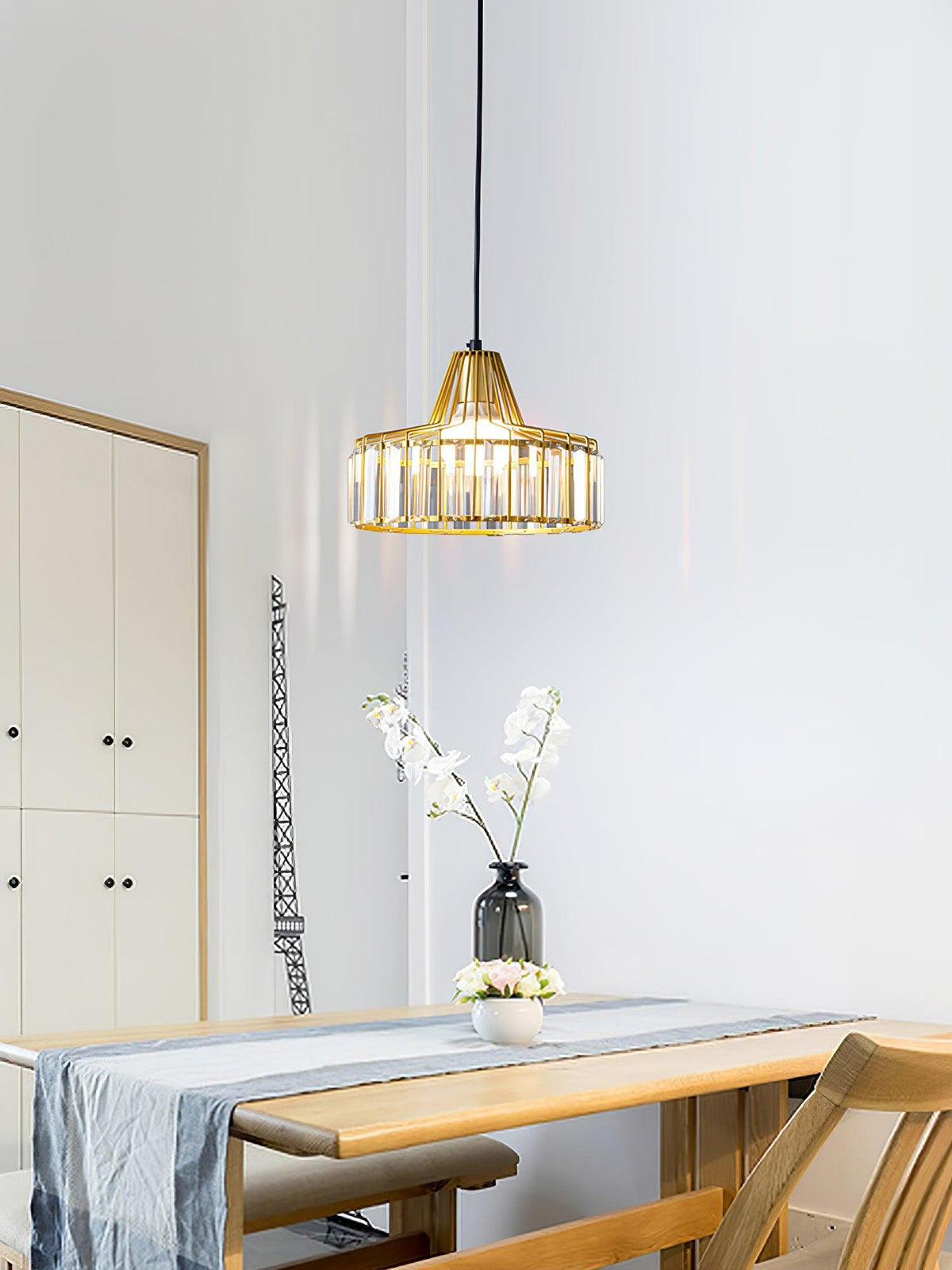 A gold crystal drum pendant from Clérachic hangs by a white cabinet, with a vase of blossom on a wooden table below.