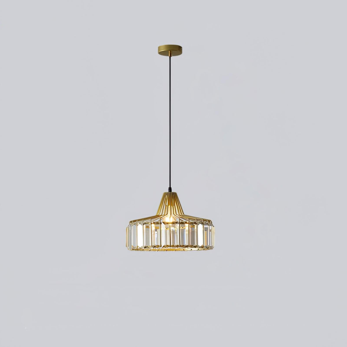 Gold crystal drum pendant from Clérachic on a gold ceiling plate, hung on a long black cable against a pale lilac-grey wall.