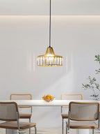 A small gold crystal drum pendant from Clérachic hangs over a white table with a bowl of oranges and cane-backed chairs.