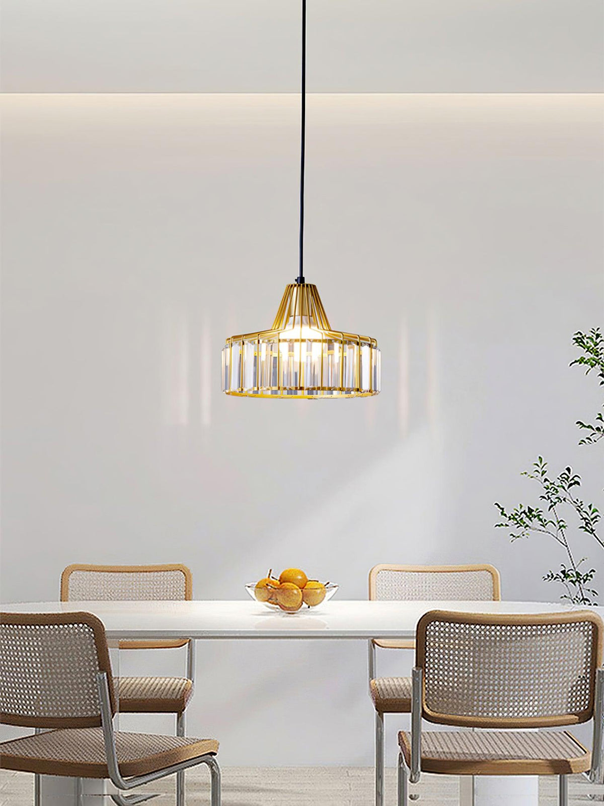 A small gold crystal drum pendant from Clérachic hangs over a white table with a bowl of oranges and cane-backed chairs.