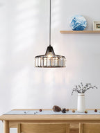 Black crystal drum pendant from Clérachic hung beside a wooden shelf and over a pale table with a vase of twigs and a pine cone.