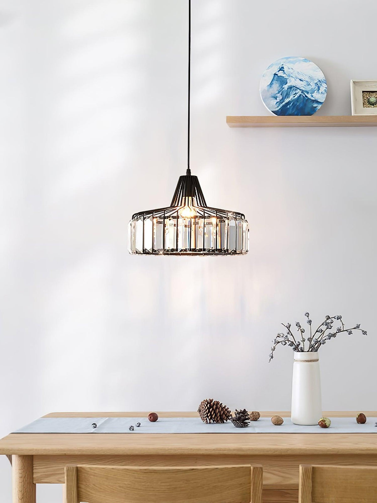 Black crystal drum pendant from Clérachic hung beside a wooden shelf and over a pale table with a vase of twigs and a pine cone.