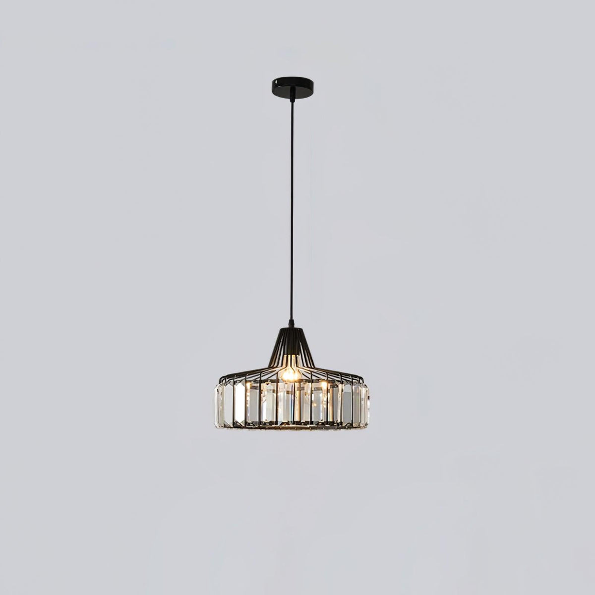 Small black crystal drum pendant from Clérachic on a black ceiling plate, hanging on a long cable against a pale grey wall.