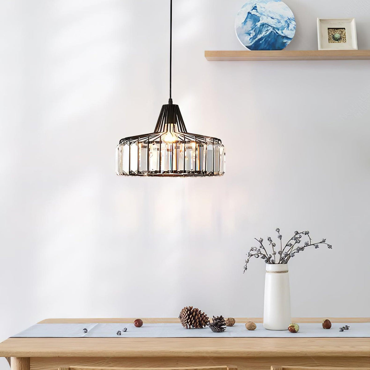 Black crystal drum pendant from Clérachic hanging in front of a pale wall, with a shelf, a vase of twigs and pine cones on the table below.