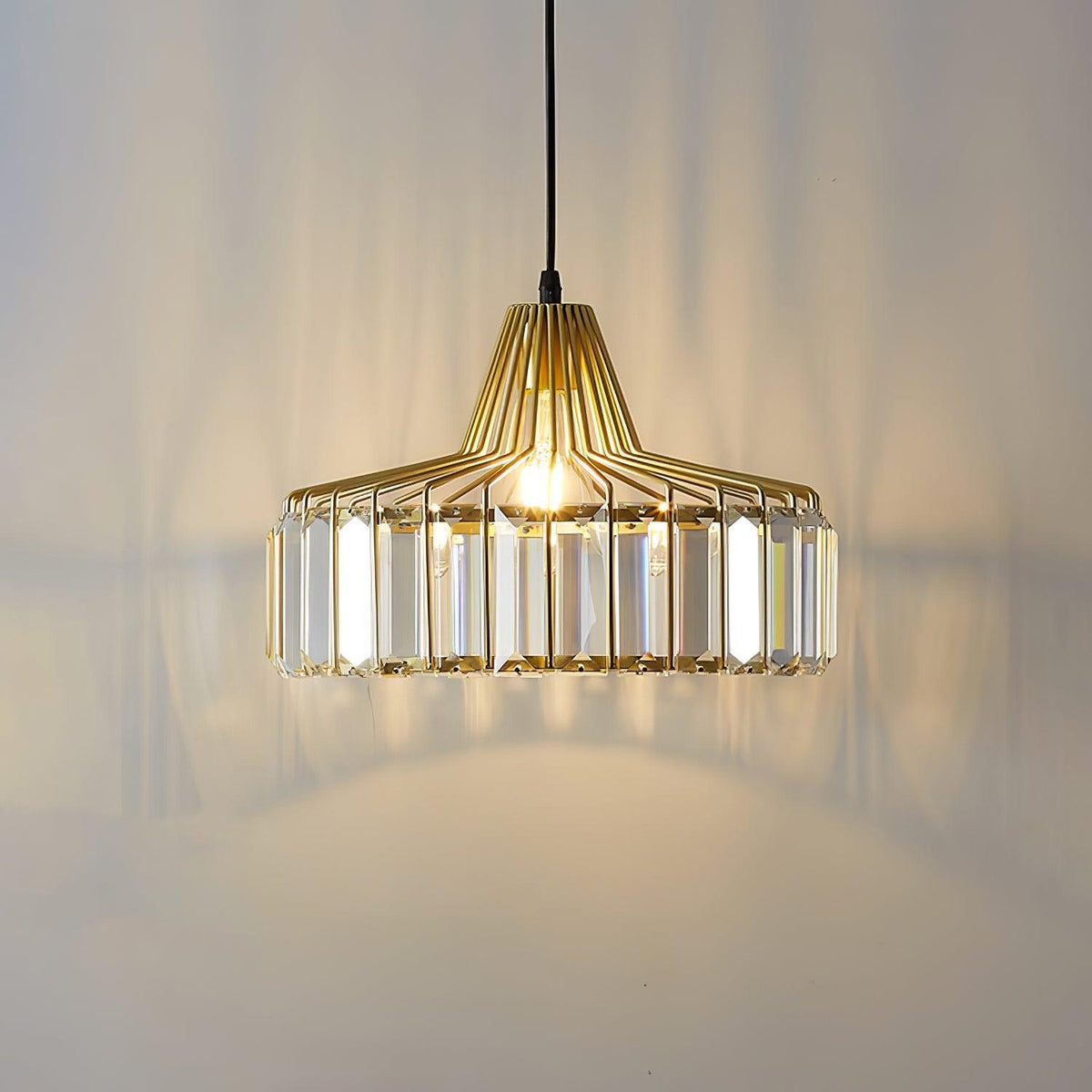 Lit gold crystal drum pendant from Clérachic, with warm light and long prism shadows across the wall behind it.