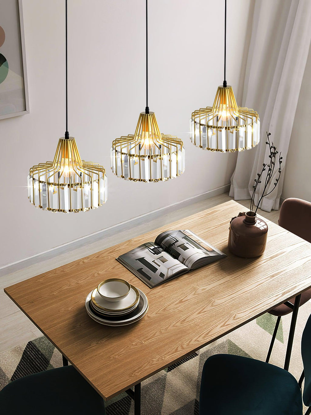 Three gold crystal drum pendants from Clérachic hang at staggered heights over a wooden table with an open magazine and a stack of bowls.