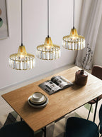 Three gold crystal drum pendants from Clérachic hang at staggered heights over a wooden table with an open magazine and a stack of bowls.