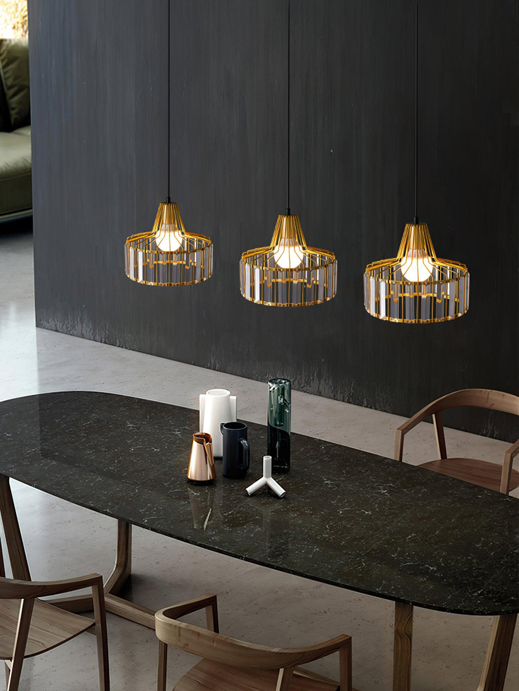 Three gold crystal drum pendants from Clérachic in a row over a dark marble-topped table with wooden chairs and a charcoal wall behind.
