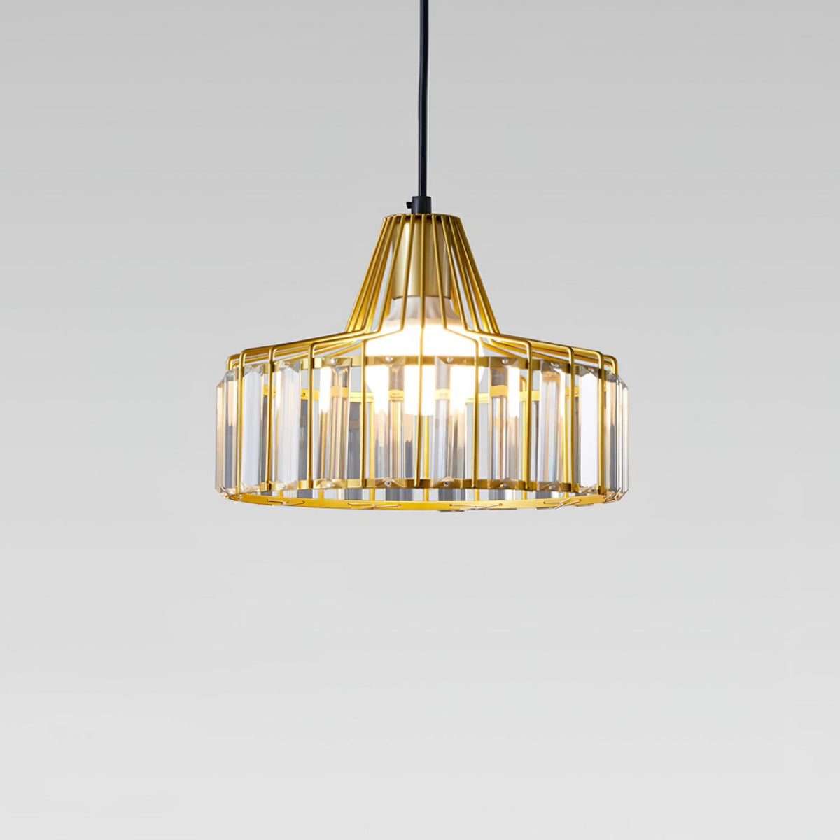 Gold wire-framed crystal drum pendant from Clérachic, with warm light glowing through the clear prisms, against a grey backdrop.