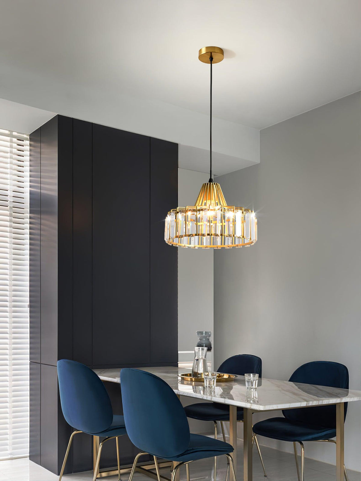 A gold crystal drum pendant from Clérachic on a brass ceiling plate hangs in a grey dining room with navy velvet chairs and a marble table.