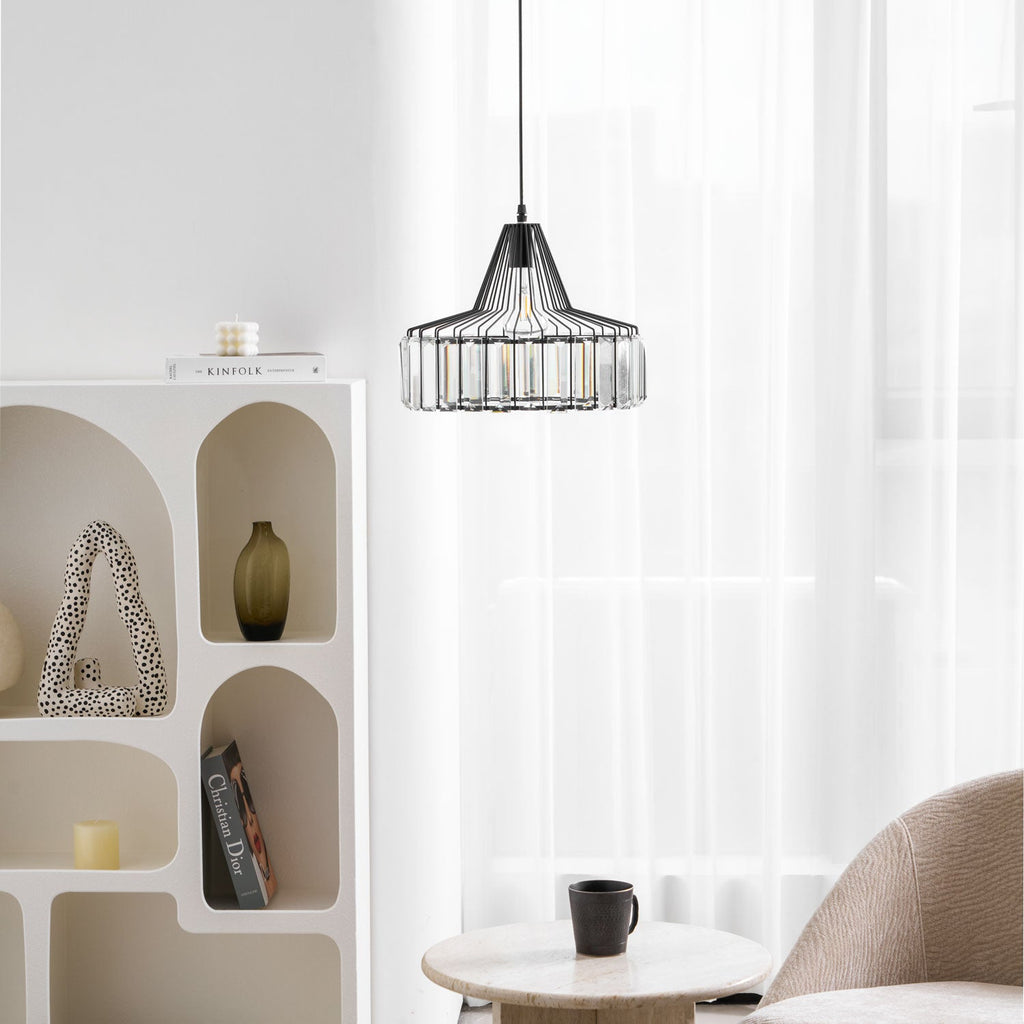 Black crystal drum pendant from Clérachic hangs by sheer curtains, with an arched white shelf unit and a small round table with a black cup.