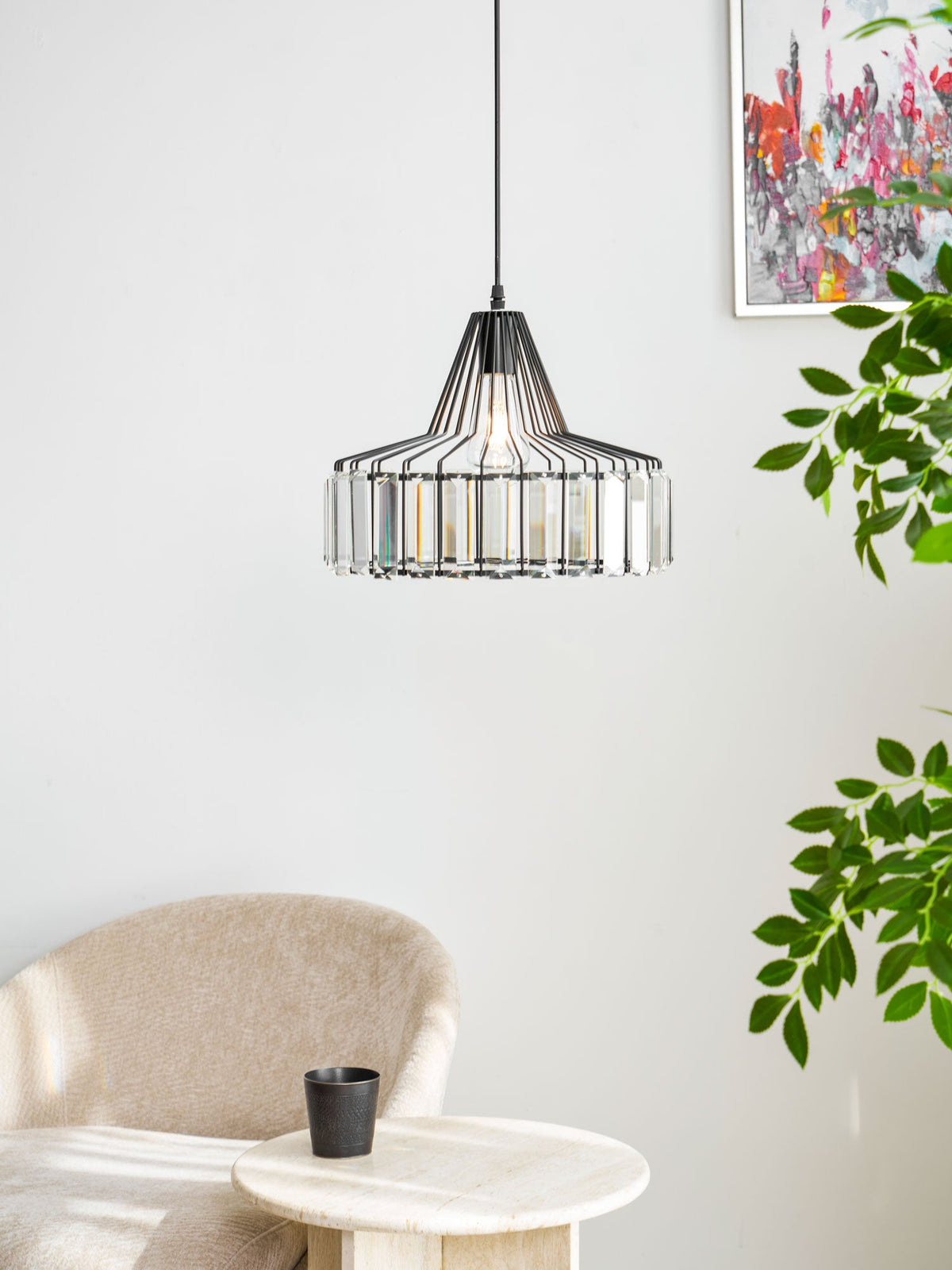 Black crystal drum pendant from Clérachic hangs against a white wall with a green plant and a beige armchair beside a round table.