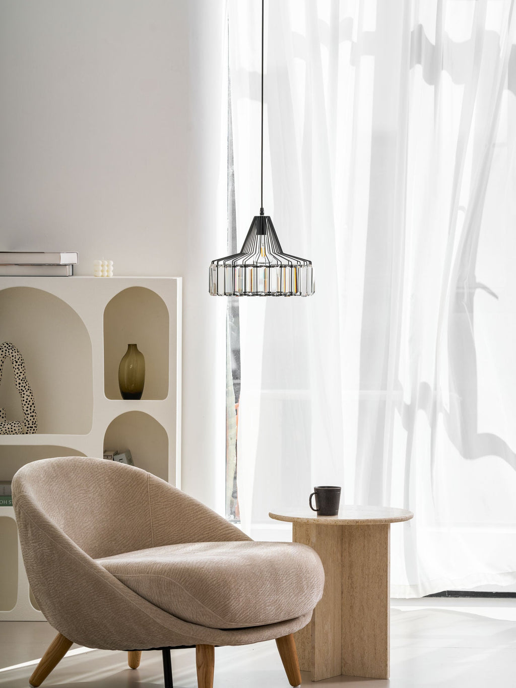 A black Clérachic crystal drum pendant hangs in front of sheer curtains, with an arched shelf unit, a beige armchair and a stone side table.