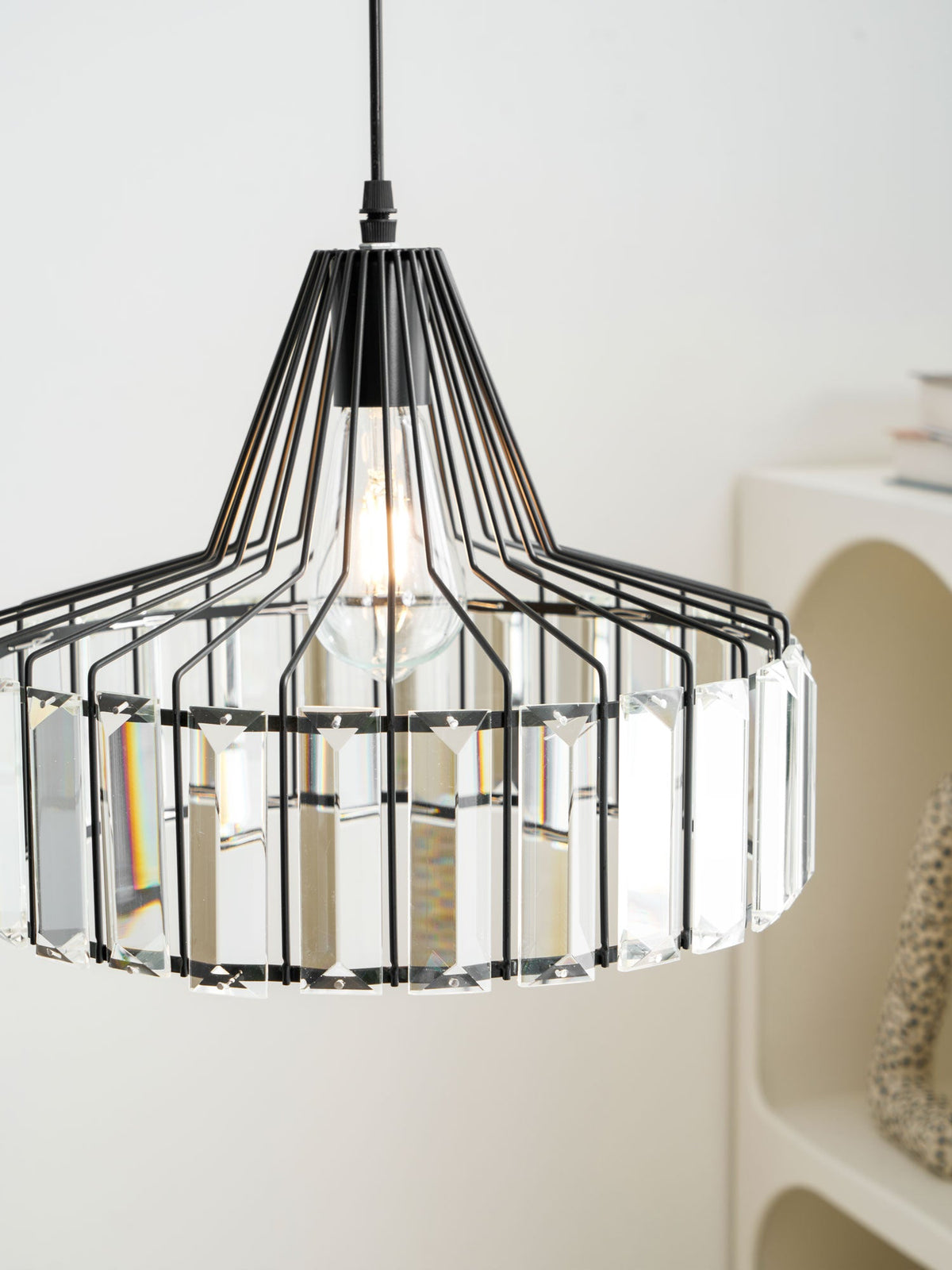 Close view of the black wire frame and clear crystal prisms on this Clérachic pendant, with an arched white shelf behind.