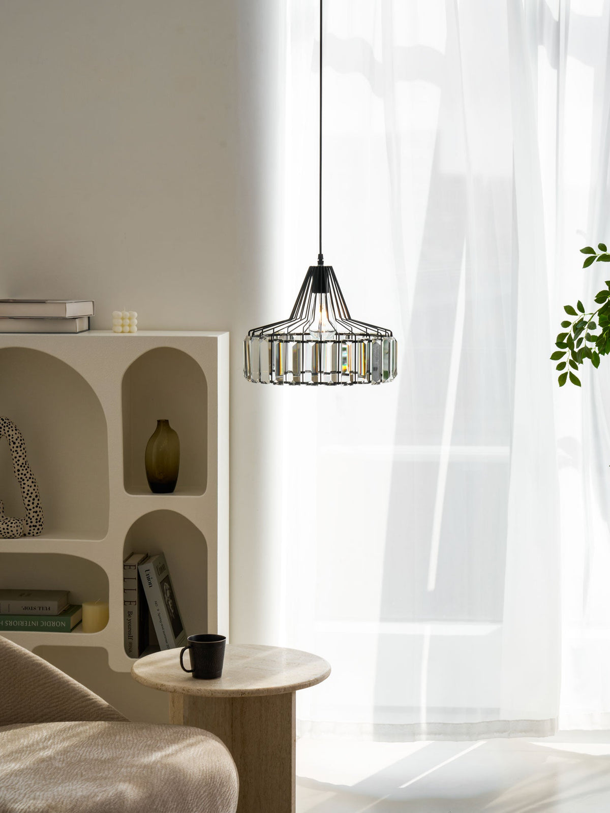 A lit black crystal drum pendant from Clérachic hangs by sheer white curtains, with a shelf unit, books and a black mug on a stone table below.
