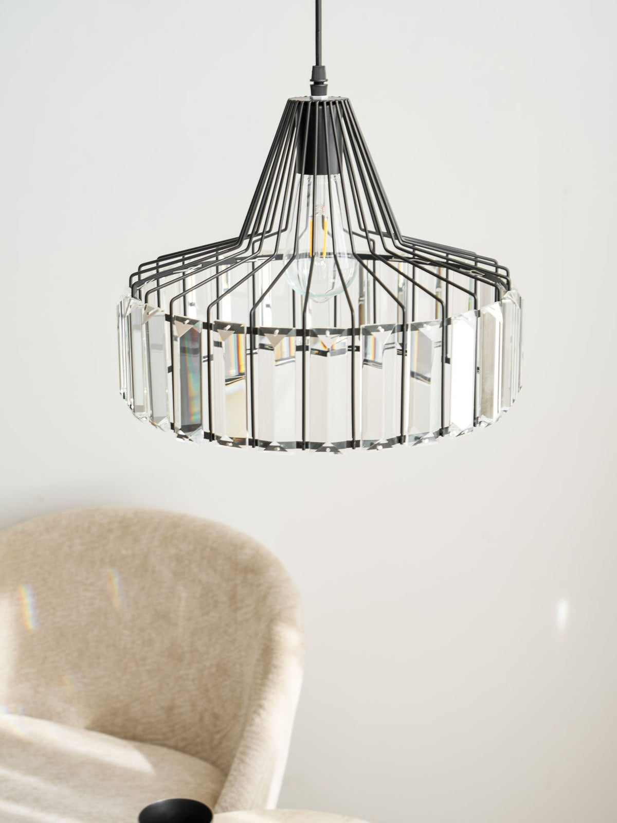 Close view of the black cage and clear prisms of this Clérachic pendant, with a beige chair and a mug softly blurred behind.