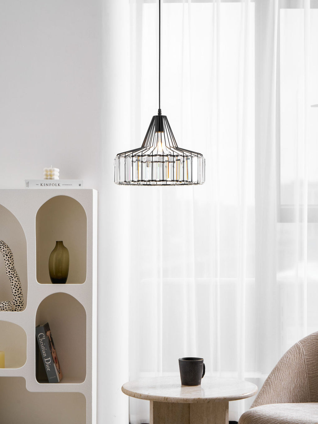 Black crystal drum pendant from Clérachic against sheer curtains, with an arched white shelf unit and a round stone table with a black cup.