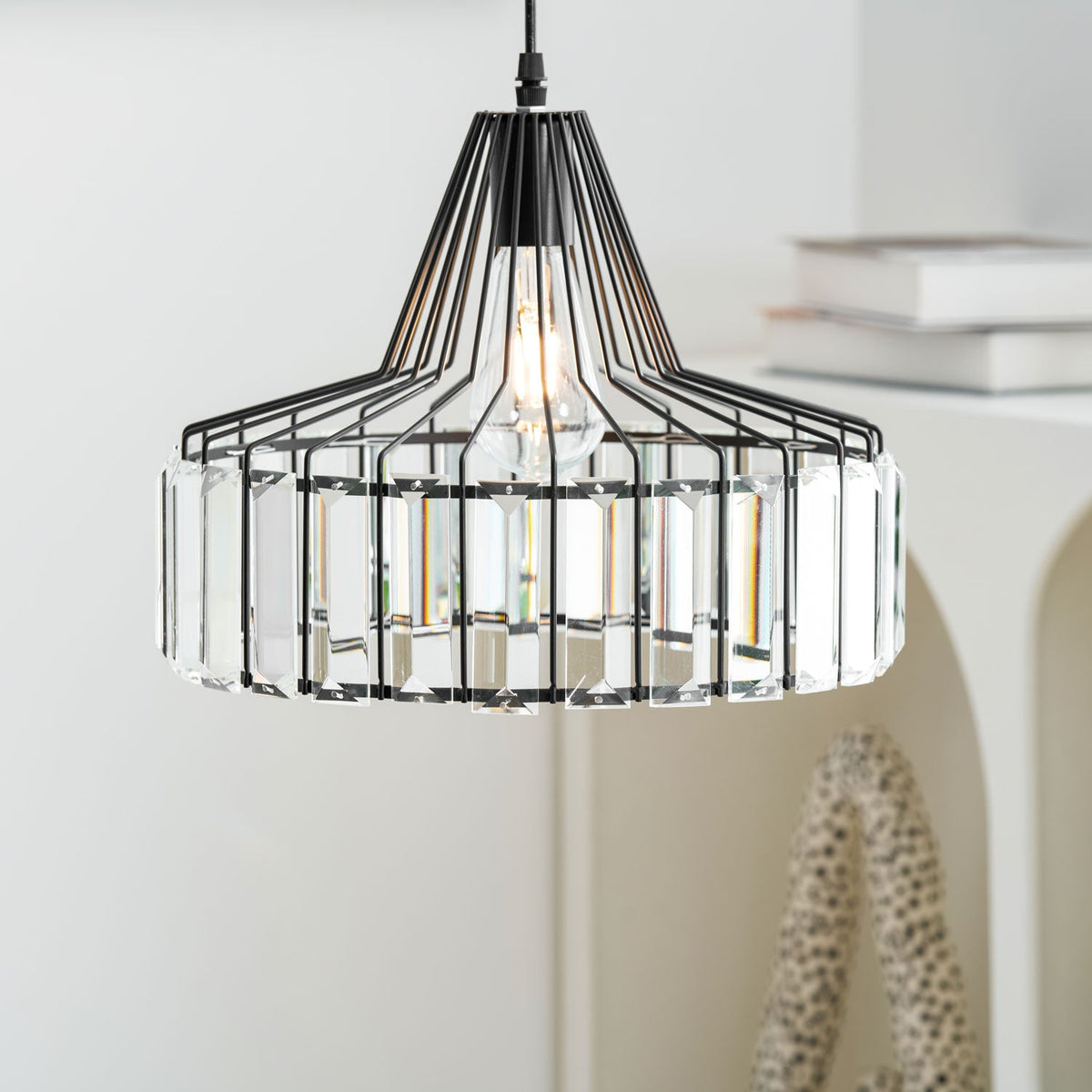 Close view of the black wire cage and clear prisms on this Clérachic pendant, with white books and a speckled ornament behind.