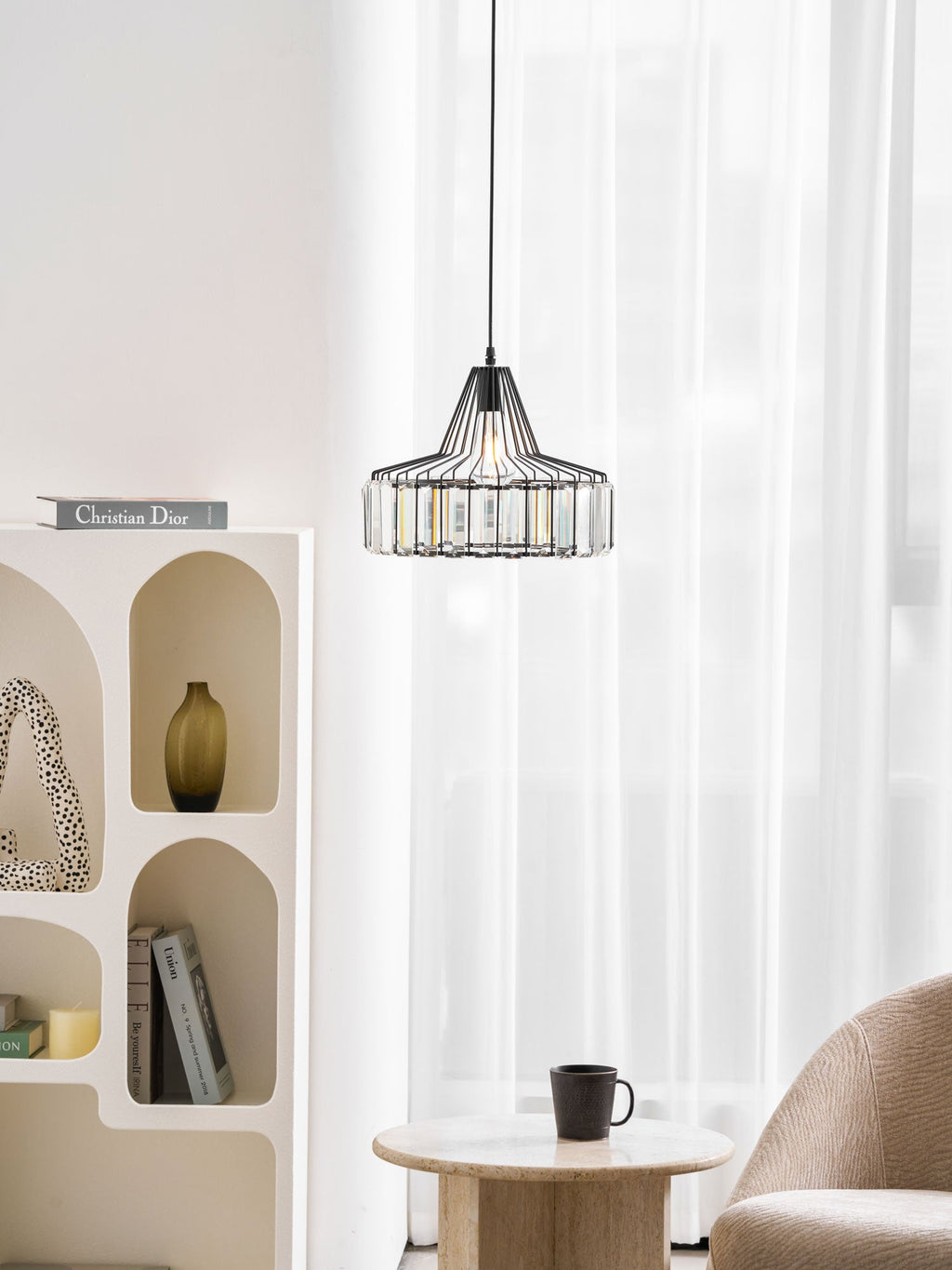 Black crystal drum pendant from Clérachic hangs by sheer curtains, above an arched white shelf unit and a table with a black cup.