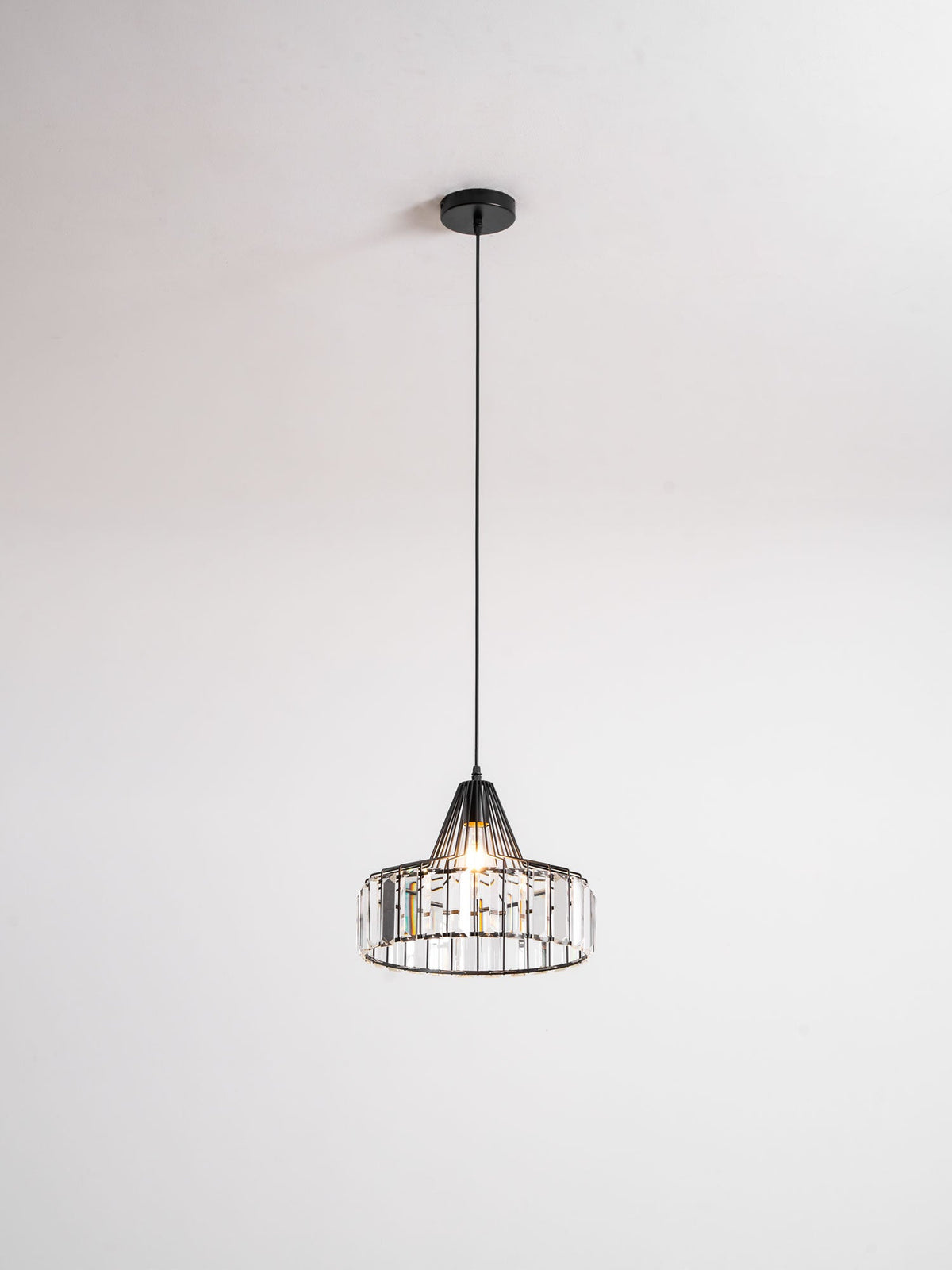 Small black crystal drum pendant from Clérachic on a round black ceiling plate, hung on a thin cable against a pale grey wall.