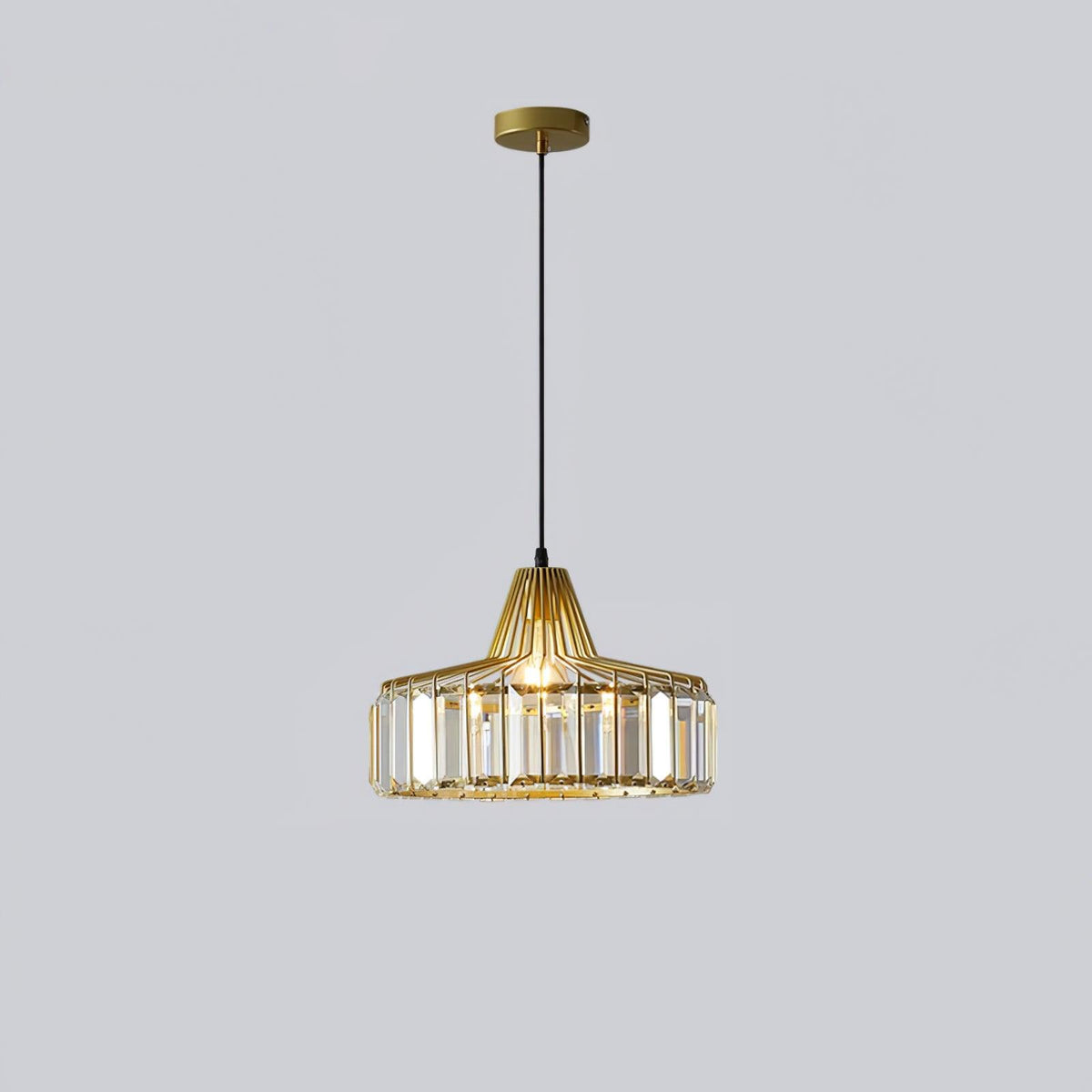 Gold crystal drum pendant from Clérachic with a gold ceiling plate, lit, hanging on a long black cable against a pale grey backdrop.