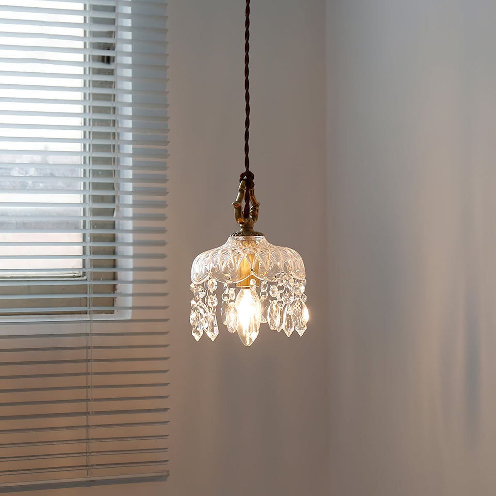 A Clérachic pendant light lit beside a window with horizontal blinds, its crystal drops catching the warm bulb glow.