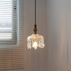 A Clérachic pendant light lit beside a window with horizontal blinds, its crystal drops catching the warm bulb glow.