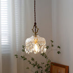 A Clérachic pendant light by a sheer-curtained window, its gold hanger and crystal drops lit warmly above a framed picture.