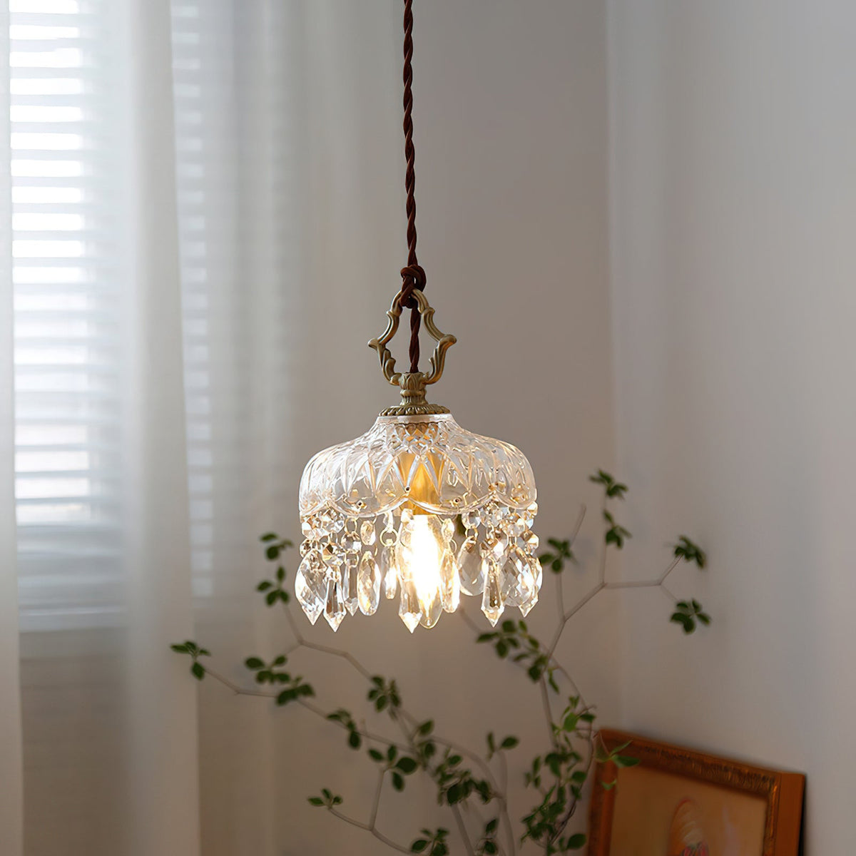 A Clérachic pendant light by a sheer-curtained window, its gold hanger and crystal drops lit warmly above a framed picture.