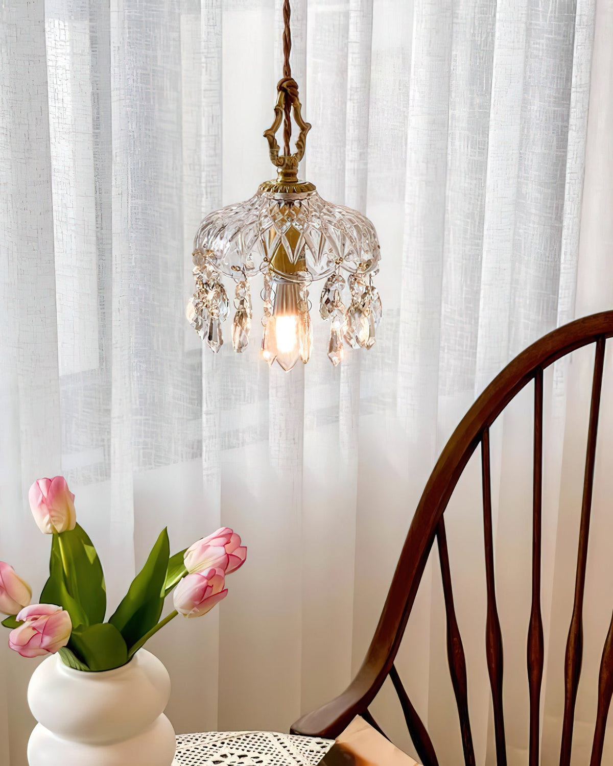 Seen through sheer white curtains, this Clérachic pendant light glows above tulips and the curved back of a wooden chair.