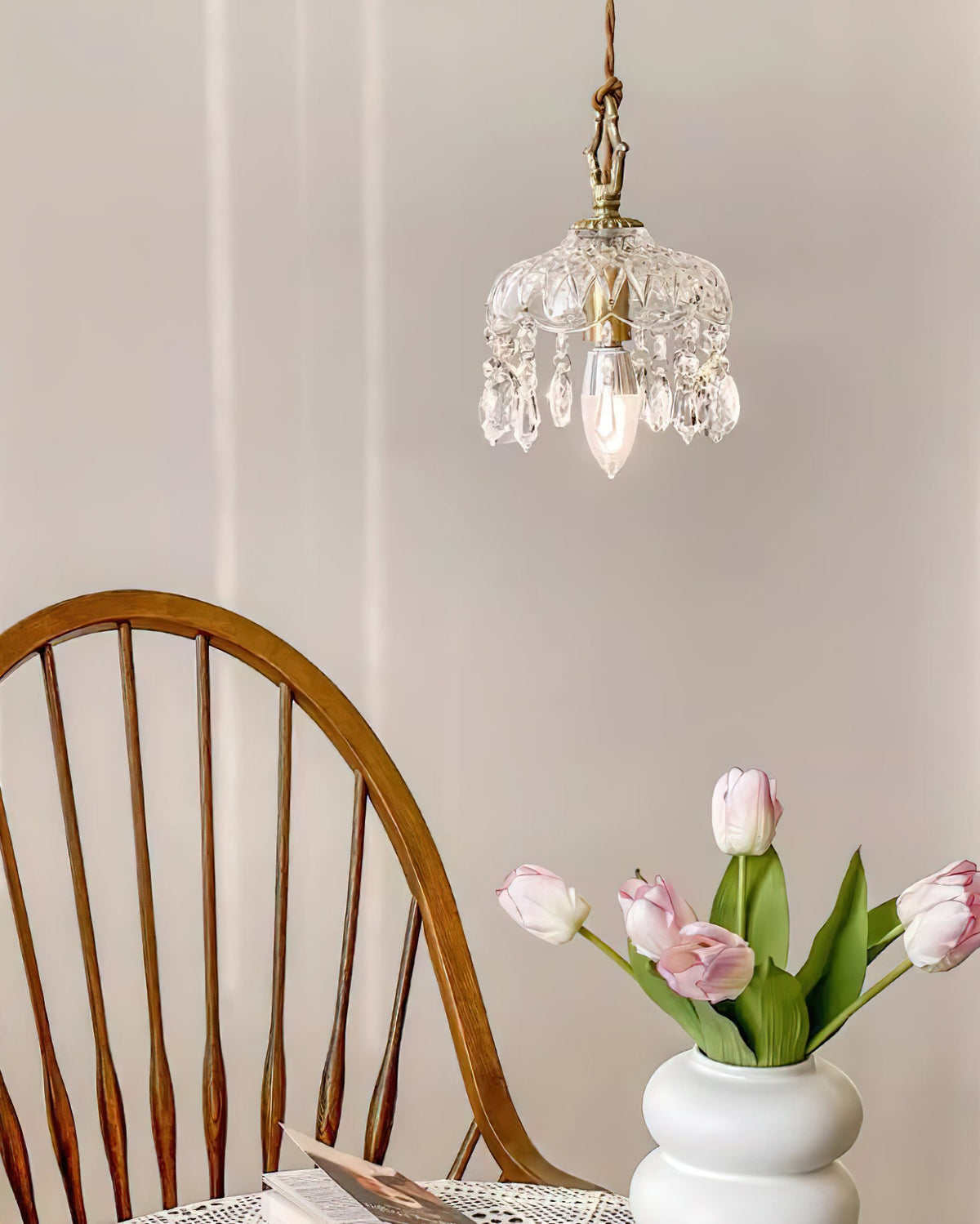 A Clérachic pendant light hangs above a spindle-back chair and a white vase of pink tulips in a soft-lit room.