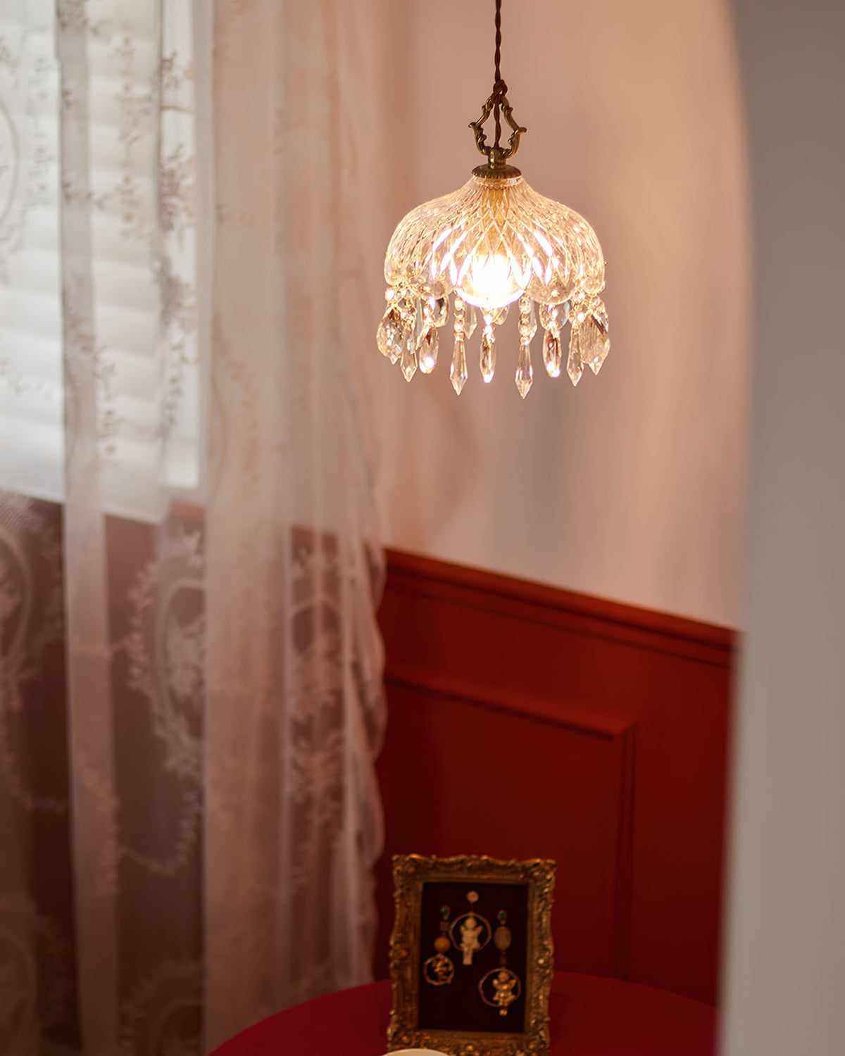 This Clérachic pendant light hangs in front of a lace curtain, with a red panelled wall and a small gilt frame behind.