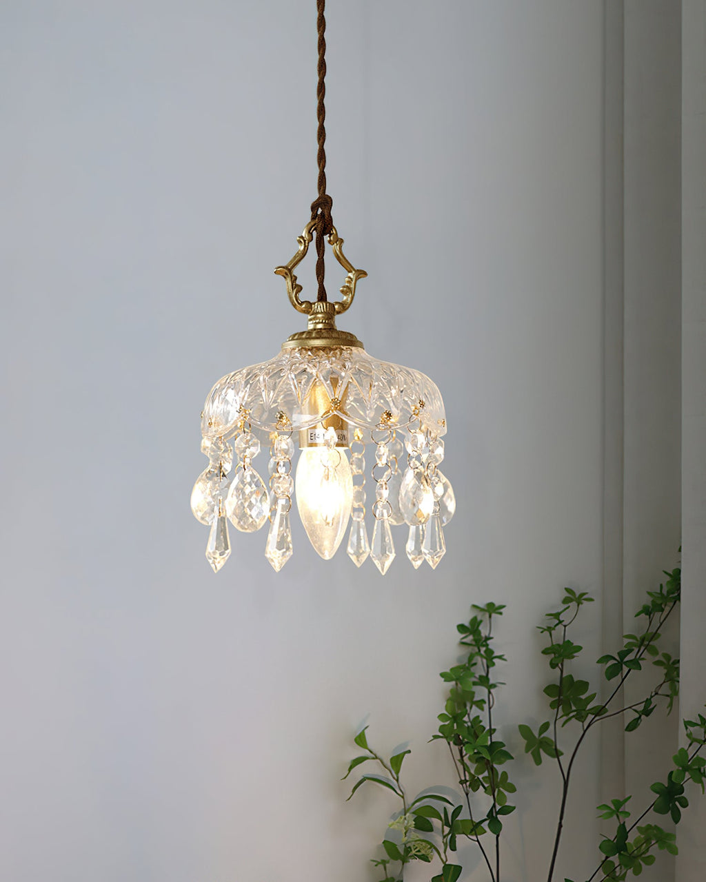 A Clérachic pendant light hangs against a pale blue-grey wall, its crystal drops lit by a candle-style bulb, with green leaves beside.