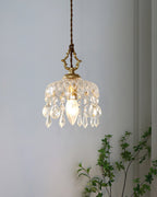 A Clérachic pendant light hangs against a pale blue-grey wall, its crystal drops lit by a candle-style bulb, with green leaves beside.