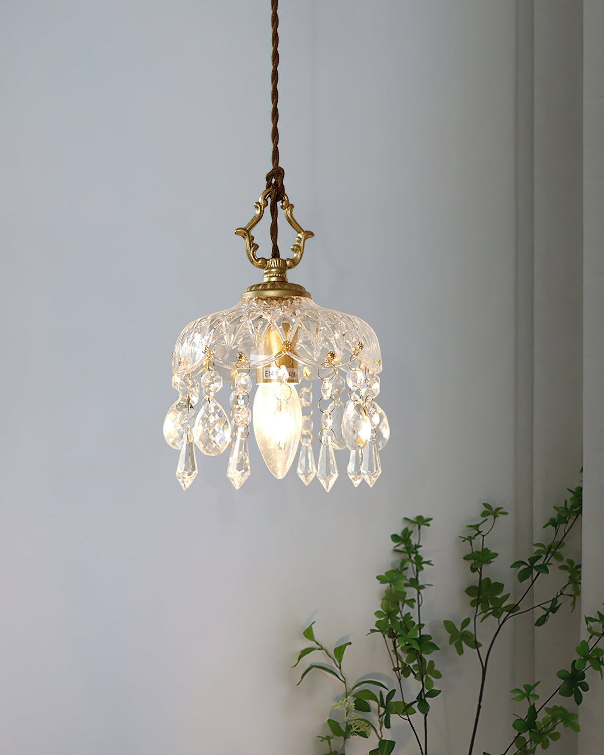 A Clérachic pendant light hangs against a pale blue-grey wall, its crystal drops lit by a candle-style bulb, with green leaves beside.